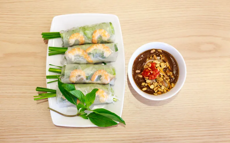 Four translucent Vietnamese summer rolls showing whole pink shrimp, rice noodles, leafy herbs and chive stems beside dark peanut-hoisin sauce topped with chopped peanuts and chile