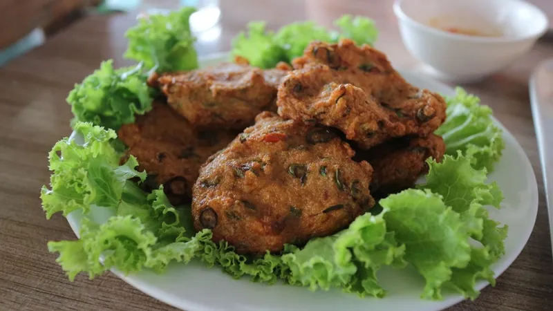 Golden Thai-style fish cakes with green bean and red pepper pieces on curly lettuce beside sweet chili dipping sauce