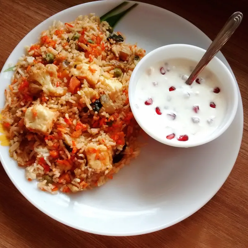 Colorful vegetable biryani with cauliflower, carrots and peas served beside pomegranate-studded raita