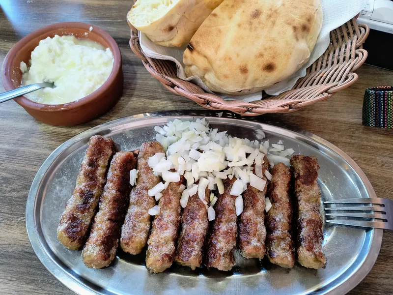 Rows of browned ćevapi topped with chopped raw onion beside round somun flatbreads and a bowl of creamy kajmak
