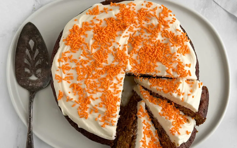 Round single-layer carrot cake with thick white cream cheese frosting and orange sprinkles, with several slices cut on a white platter