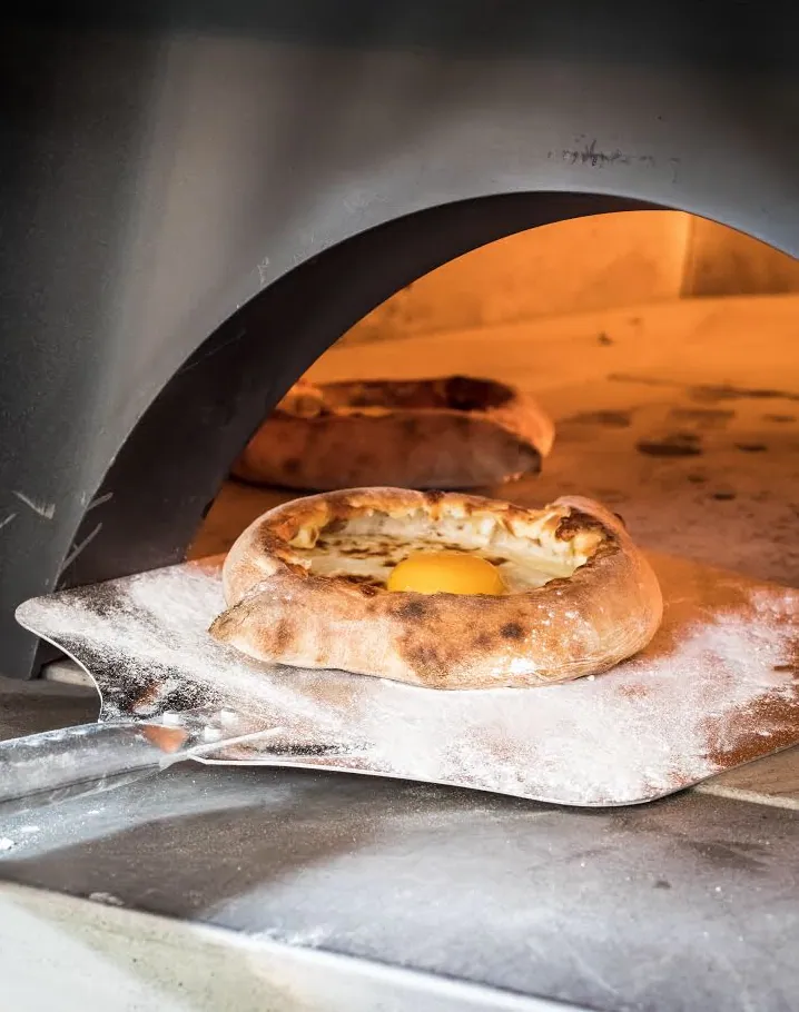 Boat-shaped Adjarian khachapuri with a browned bread rim, molten white cheese and a whole egg yolk on a metal peel at a hot oven