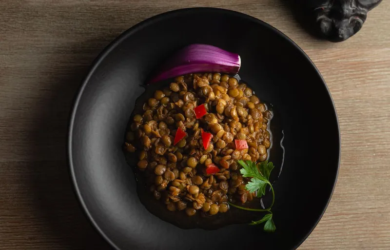 Glossy brown-green lentils with diced red pepper, a parsley sprig and a red-onion petal in a black bowl