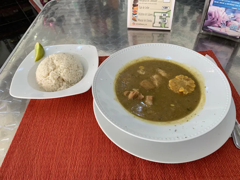 Dominican-style sancocho with pieces of chicken and pork and a corn round in a thick green-brown root-vegetable broth, served with white rice and lime
