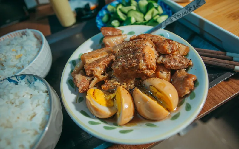 Bowl of glossy Filipino-style pork adobo made with browned pork belly pieces and halved braised eggs beside steamed white rice and sliced cucumber