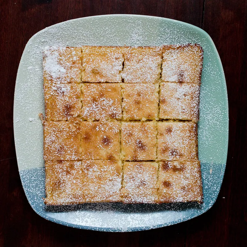 Sixteen square lemon bars with golden lemon filling and buttery shortbread crust arranged on a pale blue plate under confectioners sugar
