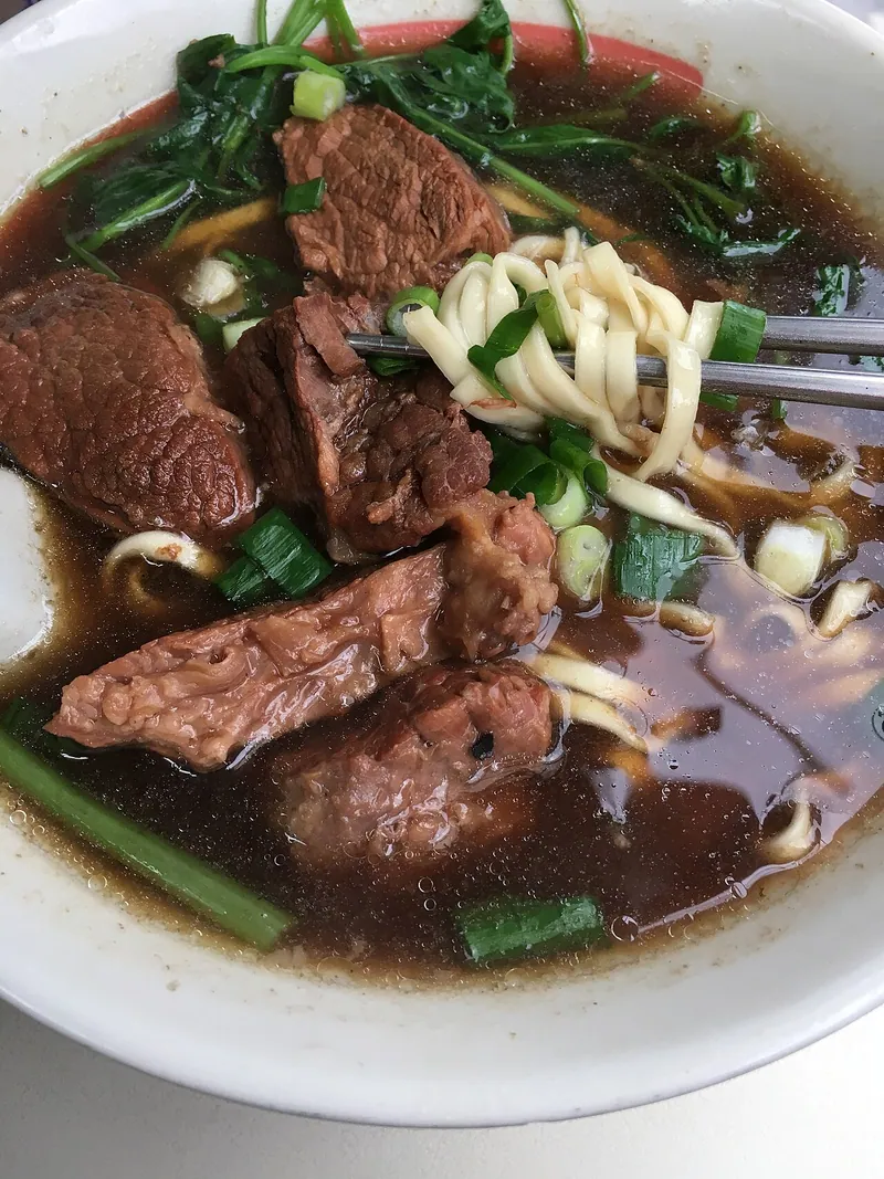 White bowl of Taiwanese-style beef noodle soup with thick wheat noodles, large braised beef chunks, dark red-brown broth, wilted leafy greens and sliced scallions