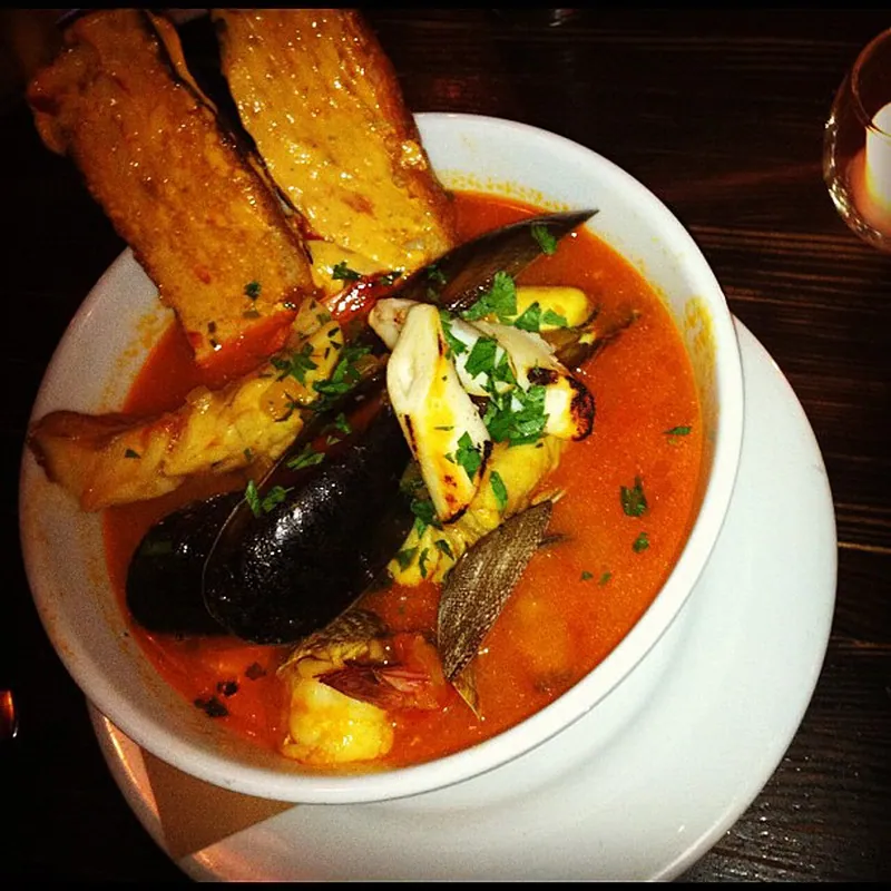 White bowl of San Francisco-style cioppino with crab pieces, dark mussel shells, clams, shrimp and white fish in red tomato broth beside two slices of grilled bread