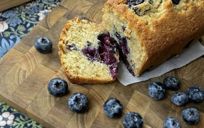 Golden blueberry loaf partially sliced on a wooden cutting board with whole fresh blueberries scattered around it
