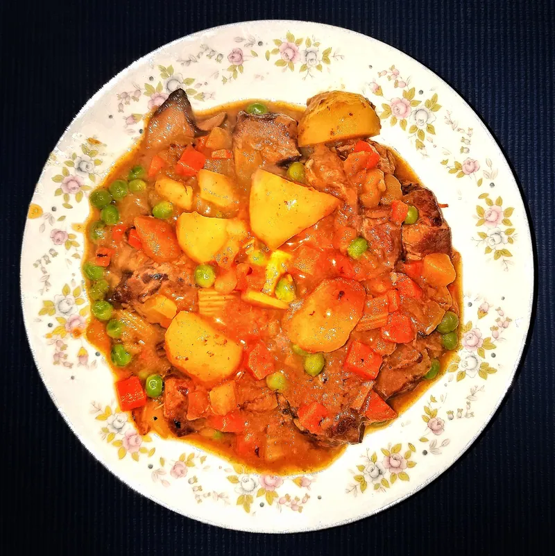 Bowl of brown beef stew with large potato pieces, carrots, mushrooms, celery and bright green peas