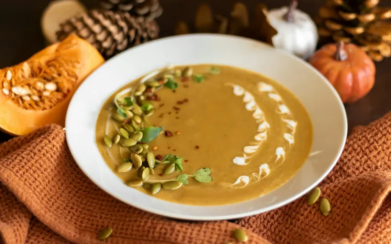 Silky orange pumpkin soup with a white crema swirl, toasted pepitas, green herbs and chile flakes in a wide white bowl