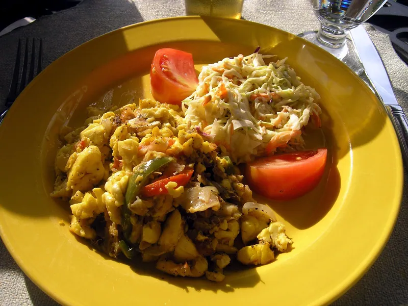 Jamaican ackee and saltfish with red and green peppers beside creamy cabbage-carrot slaw and tomato wedges on a yellow plate