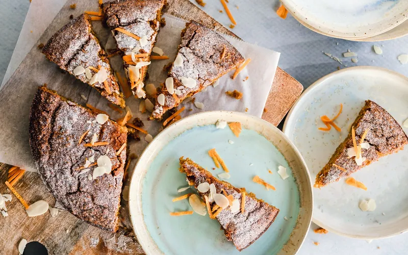 Sliced round orange-almond cake with a dark golden top, powdered sugar, sliced almonds and fine strips of orange zest on ceramic plates
