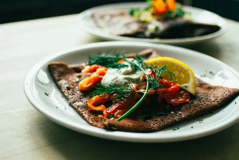 Folded brown buckwheat galettes topped with smoked salmon, crème fraîche, fresh dill and lemon wedges on white plates