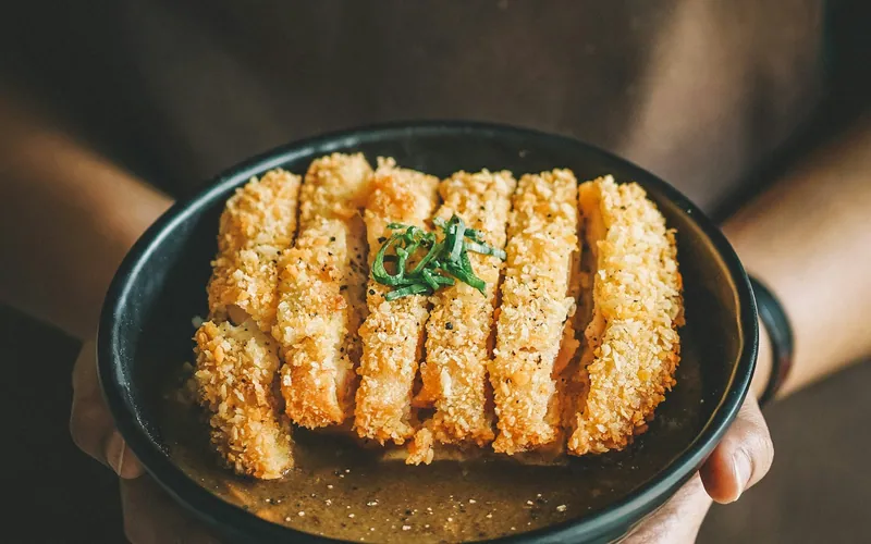 Sliced golden panko-crusted chicken katsu on a black plate with glossy brown sauce, scallions, sesame seeds, and black pepper