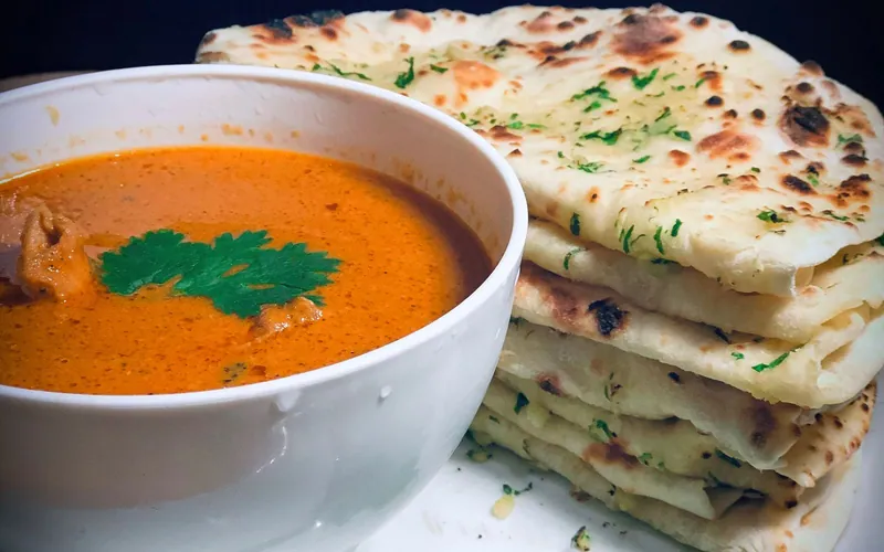 Orange butter chicken curry with visible chicken pieces and cilantro beside a stack of charred garlic naan