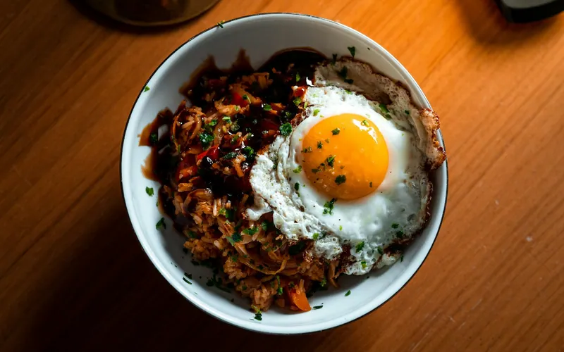 Overhead bowl of red kimchi fried rice topped with a sunny crisp-edged fried egg, chopped green scallions and a dark savory sauce drizzle