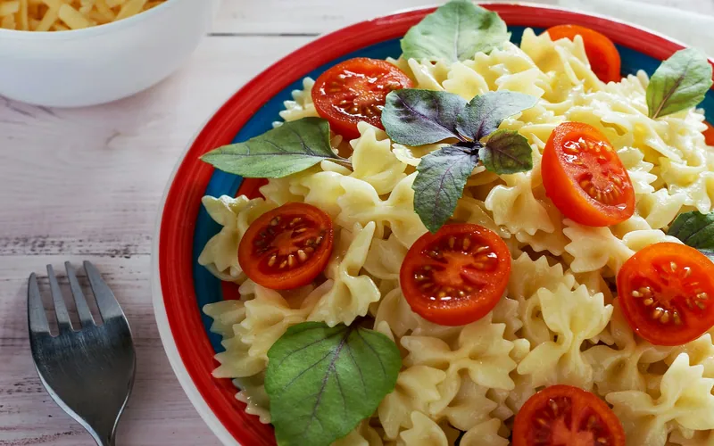 Creamy farfalle pasta with halved cherry tomatoes and fresh basil leaves on a red-rimmed plate, with grated cheese nearby
