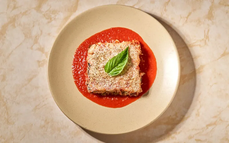 Square layered portion of baked eggplant Parmesan dusted with finely grated cheese and a basil leaf in a pool of smooth red tomato sauce