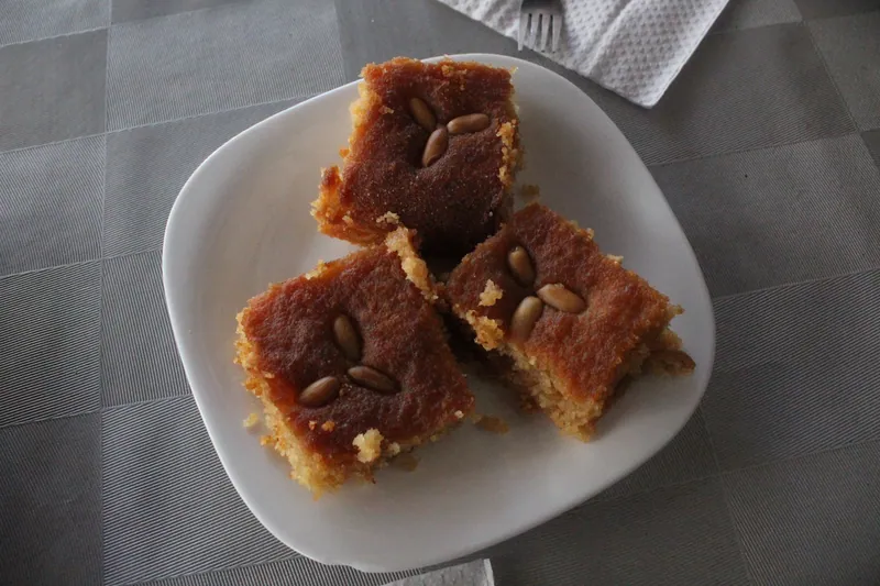 Three golden-brown squares of syrup-soaked basbousa semolina cake topped with toasted pine nuts on a white plate