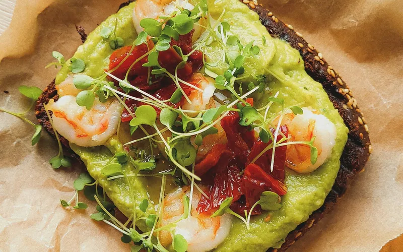 Open-faced shrimp avocado toast topped with roasted red pepper and microgreens on brown paper