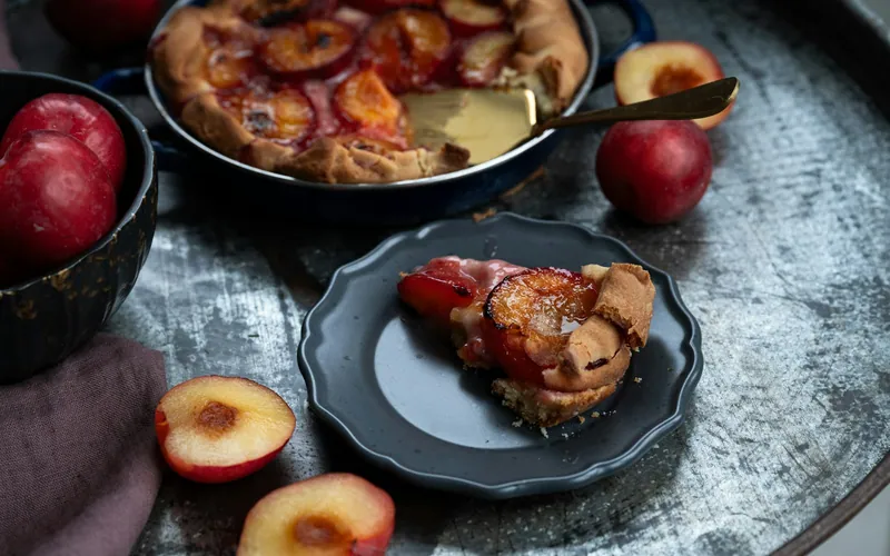 Rustic plum galette in a dark metal pan with a flaky folded crust, glossy red plum filling, a plated slice, and fresh halved plums