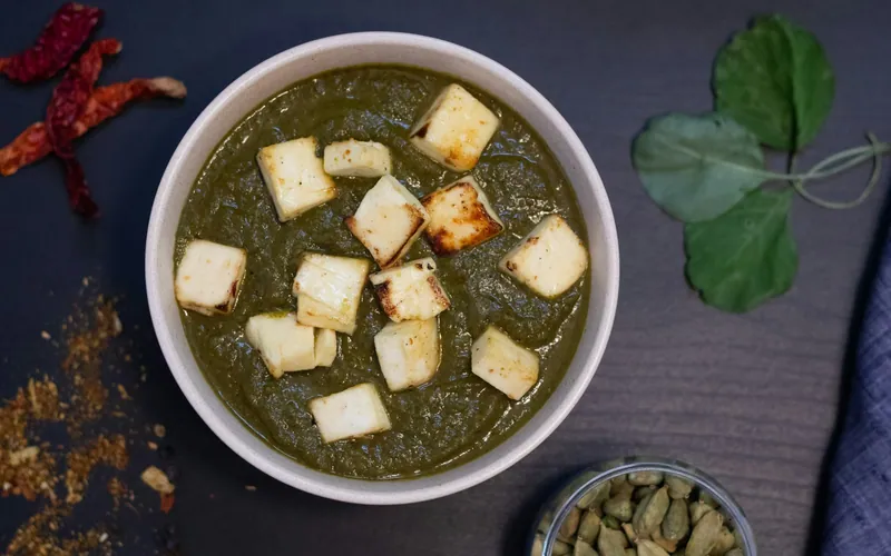Dark green palak paneer in a white bowl topped with golden-seared paneer cubes, with dried red chiles, cardamom pods, and fresh spinach leaves nearby