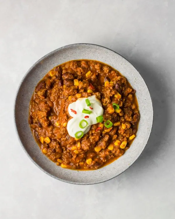 A bowl of red chili con carne with ground beef, beans, corn, sour cream, and sliced scallions
