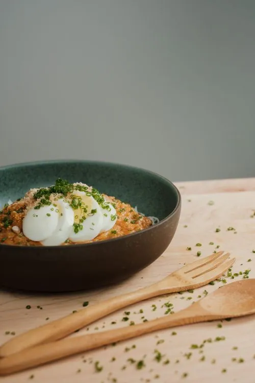 Creamy savory oat porridge topped with two poached eggs, chopped chives and a toasted sesame crumb in a dark bowl