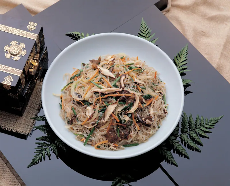 Korean japchae in a white bowl with translucent glass noodles, browned beef strips, shiitake mushrooms, spinach, carrots and scallions