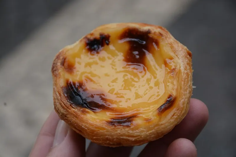 Close-up of a Portuguese custard tart with a glossy yellow center, dark caramelized blisters and many crisp golden puff-pastry layers