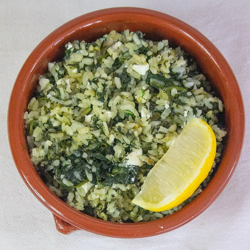 Terracotta bowl of Greek spanakorizo with visible rice and chopped spinach, flecked with fresh dill and feta beside a large lemon wedge