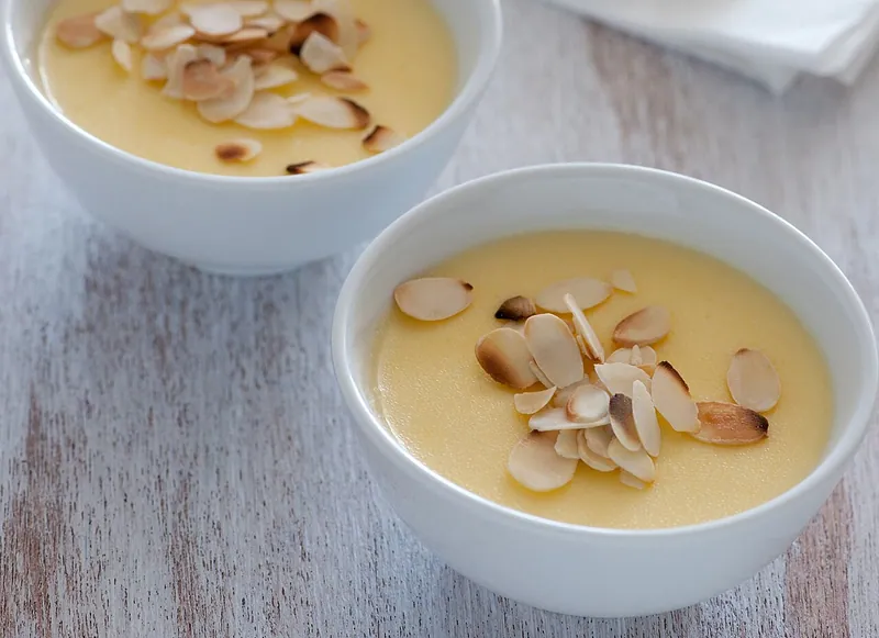 Two shallow white bowls of smooth pale-yellow lemon posset topped with toasted sliced almonds on a light wooden table