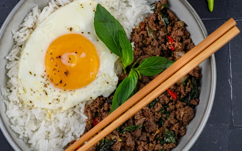 Overhead bowl of Thai basil ground beef with red chiles and fresh basil beside steamed white rice and a sunny crisp-edged fried egg