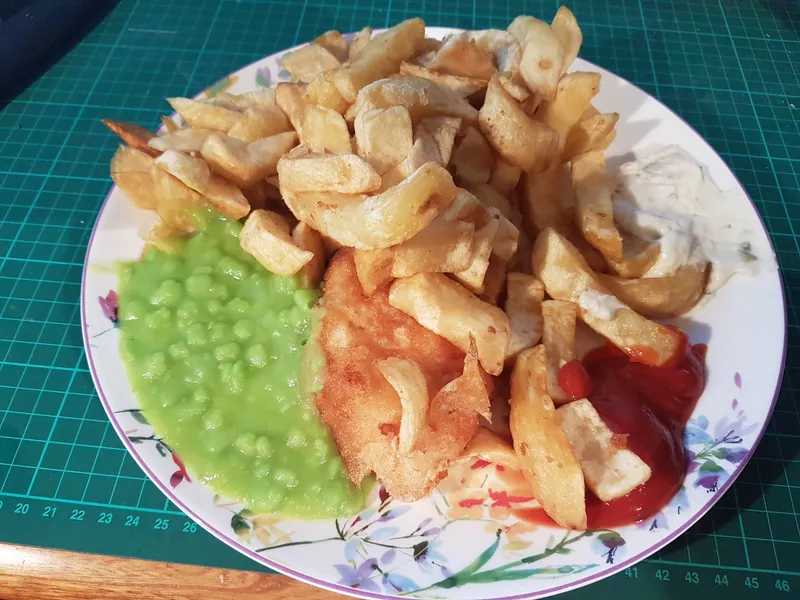 Golden battered fish with thick-cut chips, bright green mushy peas, tartar sauce and ketchup on a white plate