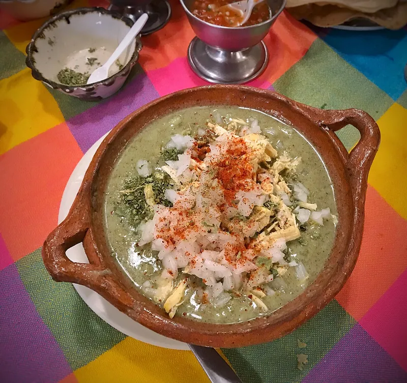 Earthenware bowl of pale-green chicken pozole with visible hominy and shredded chicken, topped with chopped white onion, dried oregano and red chile