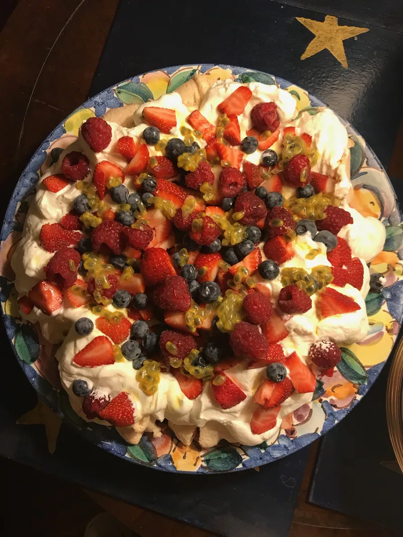 Rustic round pavlova topped with whipped cream, strawberries, raspberries, blueberries and golden passionfruit pulp