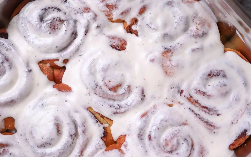 Rectangular metal pan filled edge to edge with tall golden cinnamon rolls covered generously in white vanilla cream cheese frosting