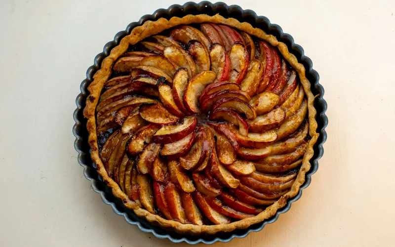 Round cinnamon-apple tart in a dark fluted pan with overlapping red-skinned apple slices, a golden buttery crust, caramelized edges, and a glossy apricot finish