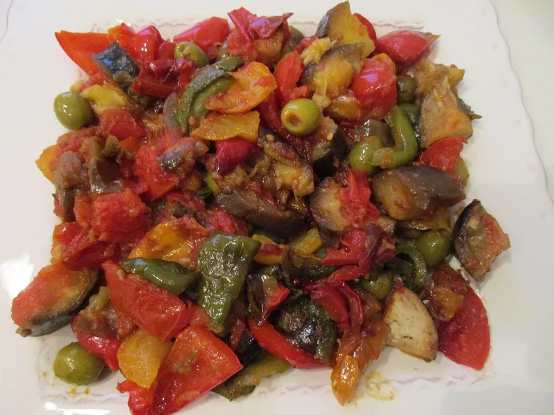 White platter of chunky Provençal ratatouille with red tomatoes, purple eggplant, green zucchini, peppers and whole green olives