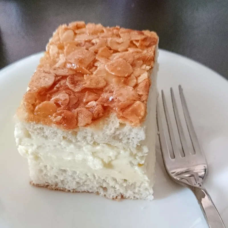 Slice of German Bienenstich with two tender yeast-cake layers, a thick pale vanilla-cream center and a glossy sliced-almond caramel top
