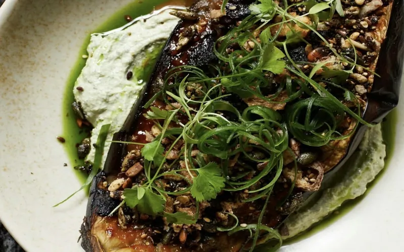 Charred miso-glazed eggplant half on pale green herb tahini with curled scallions, cilantro, herb oil, and toasted seed crunch