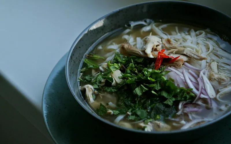 Dark bowl of clear chicken pho with flat rice noodles, shredded chicken, sliced red onion, red chile, cilantro and mint