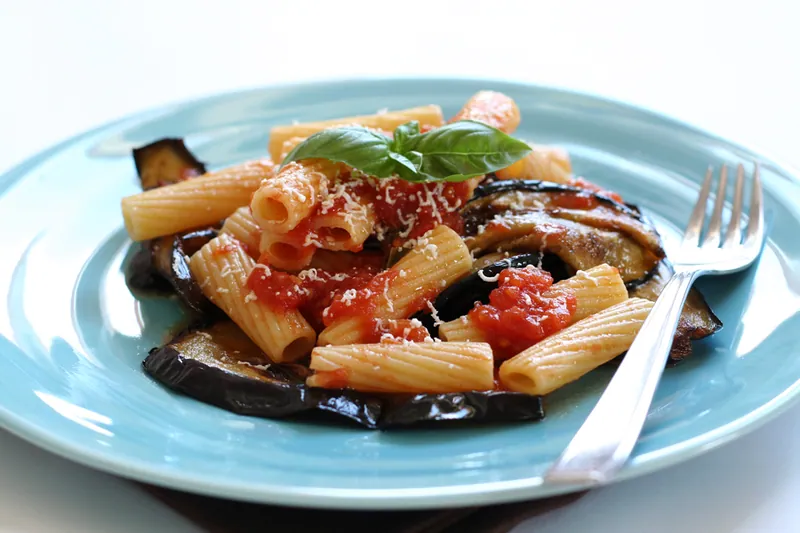 Rigatoni in chunky tomato sauce with browned eggplant slices, grated ricotta salata and fresh basil on a blue plate