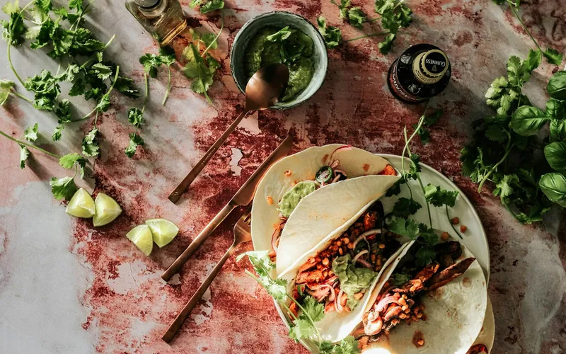 Soft flour tortillas filled with roasted sweet potato, mushrooms, corn, red onion and cilantro beside a bowl of green guacamole and lime wedges