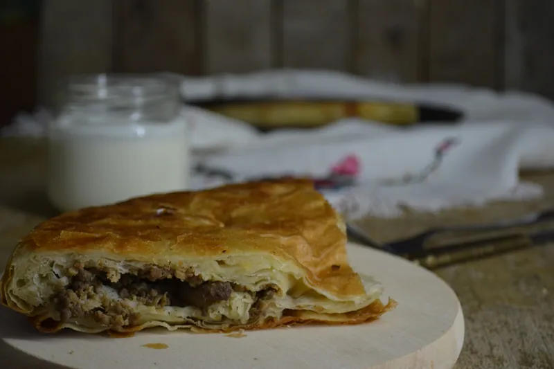 Cut square of Balkan-style meat burek showing browned minced-meat filling between crisp golden phyllo layers with yogurt behind it