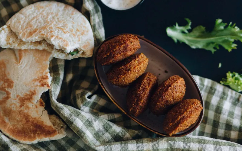 Five deep-golden oval falafel on a brown platter beside torn warm pita, leafy greens and a small bowl of pale lemon-tahini sauce