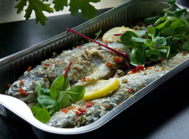 Two roasted whole trout with lemon wedges, chopped chile, garlic, parsley, and fresh greens in a metal roasting pan