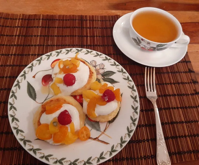 Golden round syrniki topped with sour cream, raspberries and candied mango and kumquat on a floral plate